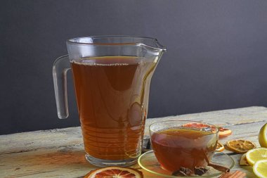Glass cup of black tea with cinnamon sticks, anise star, lemon and dried fruit on white wooden table background. Jug of black tea. Large copy space.