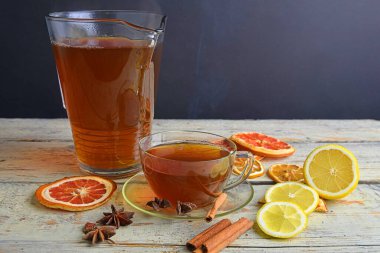 Glass cup of black tea with cinnamon sticks, anise star, lemon and dried fruit on white wooden table background. Jug of black tea. Large copy space. Black background empty space for text.