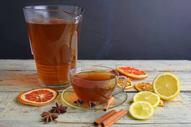 Warm cup of tea and glass jug on wooden rustic table. Cinnamon sticks, star anise, lemon and dried fruit. Black background with copy space. Still life concept.