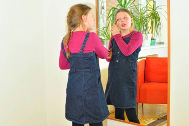 Güzellik ve moda konsepti. Güzel yanaklı çocuk gülümsüyor ve içeride dudak boyuyor. Küçük kız odada aynanın önünde ruj ya da rimel tutuyor. Çocuk dudak boyamaya ve makyaj yapmaya çalışıyor.