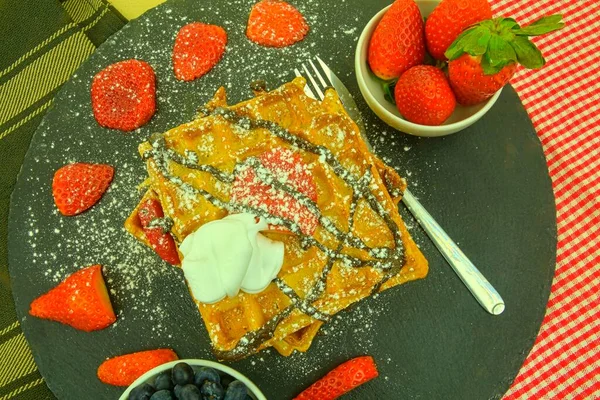 Waffle Belgia persegi dengan buah beri dan gula icing di atas piring hitam. Wafel manis manis yang lezat. Cokelat meleleh dan krim kocok pada wafel. Tampilan atas — Stok Foto