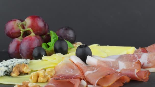 Assorted cheeses on black slate board. Brie cheese, cheddar slices, walnuts, grapes, olives and Schwarzwald ham. Still life food. Copy space — Stock Video