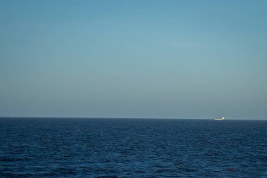 Mavi sulu deniz manzarası ve beyaz köpüklü dalgalar. Ufukta bir nakliye gemisi görülebilir..