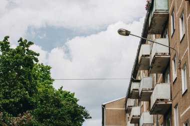 Yanında balkonları olan eski bir apartman dairesi. Yanında gölge yaratan yeşil yapraklı bir ağaç var. Beyaz kümülüs bulutlarıyla sıcak bir yaz günü.