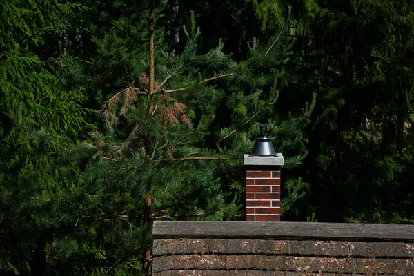 Red brick chimney for a sauna house with a wooden tile roof covered with tree needles and leaves.