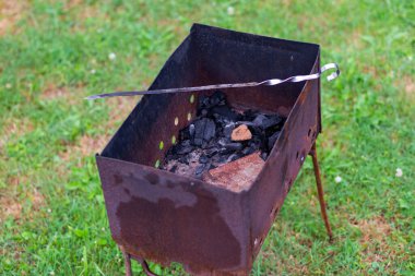 rusted metal grill with metal legs in green grass with a silver skewer across it. Black unburnt coals remained in the grill.