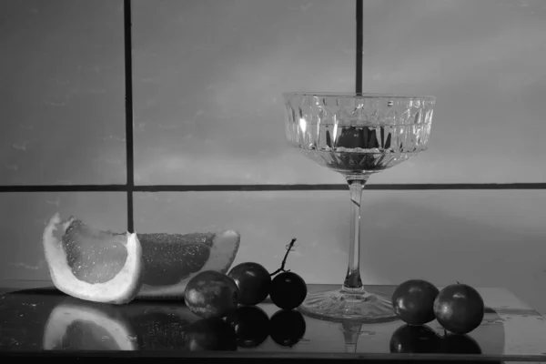 Crystal glass for cocktails on a glass surface. Fruits - grapes, grapefruit next to the glass. Abstract background.