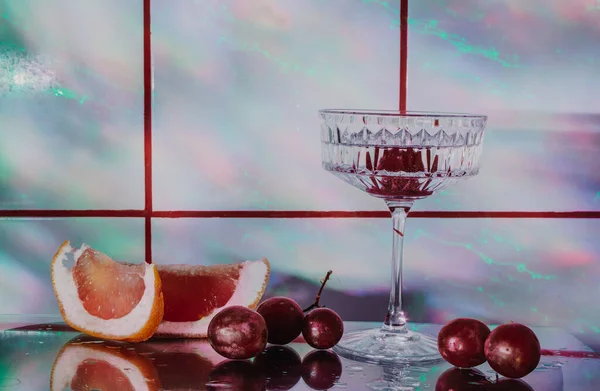 Crystal glass for cocktails on a glass surface. Fruits - grapes, grapefruit next to the glass. Abstract background.