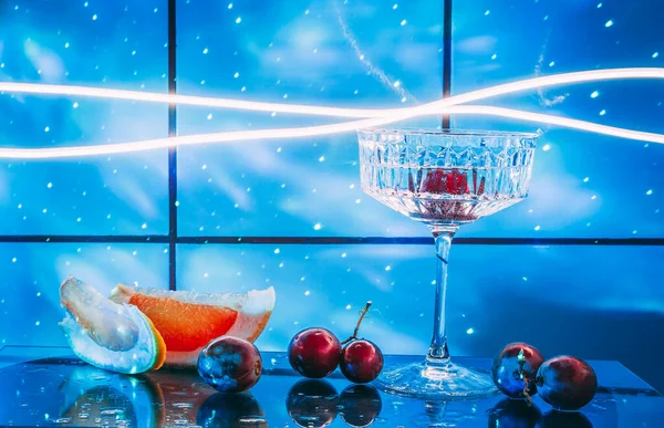 Crystal glass for cocktails on a glass surface. Fruits - grapes, grapefruit next to the glass. Abstract background.