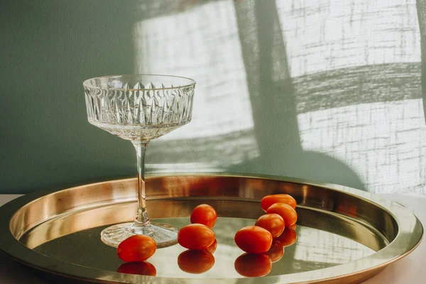 beautiful crystal glass with red tomatoes stands on a gilded metal tray illuminated by sunlight, casting a shadow on a white wall.