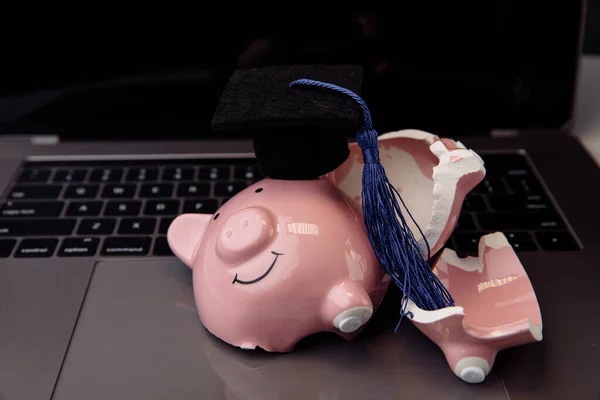 Broken pink piggy bank in cap on keyboard. College, graduate, diploma concept