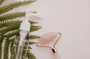 Pembe kuvars yüz masajı silindiri, şişenin özü ve pembe masanın üzerinde eğrelti otu yaprağı.
