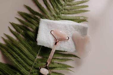 Pembe gua sha ve kuvars yüz masajı merdiveni yapraklı bir havlu üzerinde. Güzellik ve bencillik konsepti. Üst görünüm