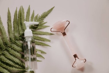 Pembe kuvars yüz masajı silindiri, şişenin özü ve pembe arka planda eğreltiotu yaprağı.