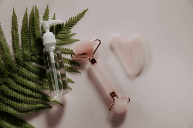Gua sha masaj kazıyıcısı ve pembe kuvars yüz masajı silindiri, şişenin içindeki özü ve pembe masanın üzerindeki eğreltiotu yaprağı.