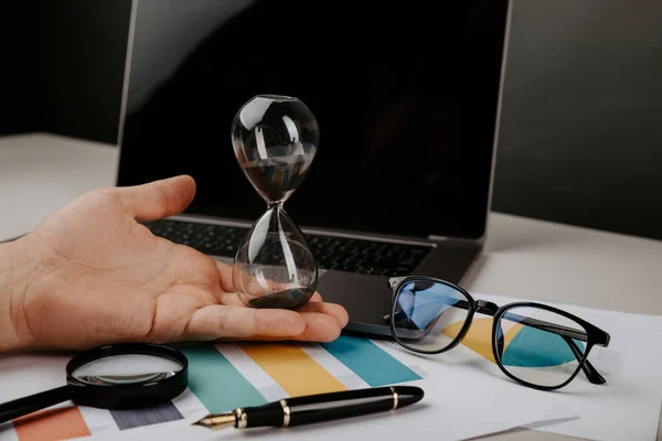 Reloj de arena en una mano de hombre de negocios, portátil y lupa en el escritorio —  Fotos de Stock