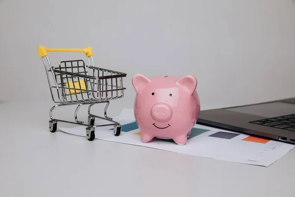 Carrinho de compras com banco porquinho e laptop em uma mesa. Conceito de compras on-line, compra e negócios — Fotografia de Stock