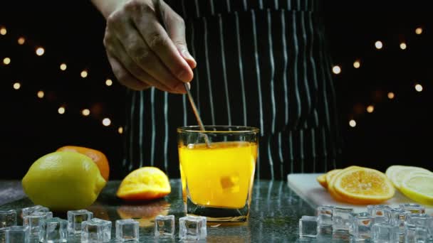 男はテーブルの上でオレンジジュースグラスに氷をかき混ぜてバーテンダーはバーでパーティーで飲むためのフルーツカクテルやフレッシュジュースを準備します — ストック動画