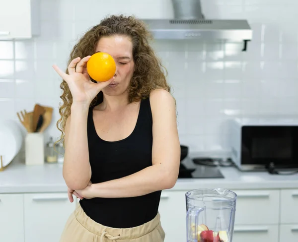 smart vegetarian woman happy funny moment by hiding and closing eye behind orange while cooking fruit juice in kitchen. beautiful caucasian woman cooking healthy diet fruit or vegetable in kitchen