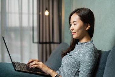 Bilgisayarla çalışan Asyalı iş kadını, rahat giyin, kanepeye otur, dizüstü bilgisayara bak, pencere arkasındaki boşluğu kopyala. Genç bir kadın ofis ve fotokopi alanında çalışıyor.