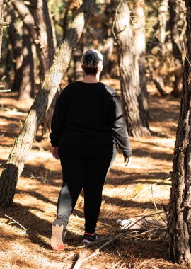 Kısa saçlı, siyah eşofmanlı, tanınmayan bir kadın sonbahar ormanında yerde yapraklarla yürüyor.