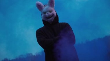 scary evil maniac in a rabbit mask looks at the camera and holds colored smoke