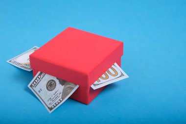Dolar banknotları mavi arka planda kırmızı bir hediye kutusuna sıkışmış.