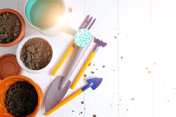 Çiçek saksıları, sulama kabı ve bahçe aletleri beyaz tezgahın üzerinde.