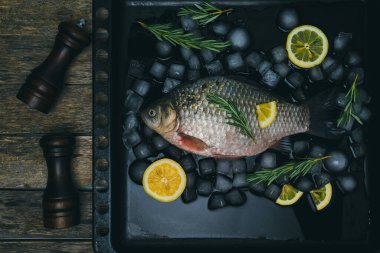 Pişirme balığı, buz küplü çiğ balık, limon dilimleri ve biberiye sprigleri. Ahşap arka planda baharat kavanozları.