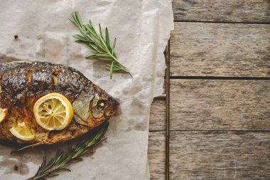Ahşap bir arka planda baharat, limon ve biberiye ile parşömen kağıdında pişmiş balık.