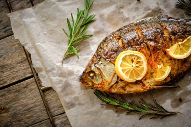 Ahşap bir arka planda baharat, limon ve biberiye ile parşömen kağıdında pişmiş balık.