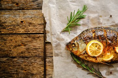 Ahşap bir arka planda baharat, limon ve biberiye ile parşömen kağıdında pişmiş balık.