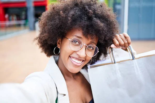 Happy young afro woman taking a selfie with her mobile phone while shopping. She is holding the bags with her hands while smiling.