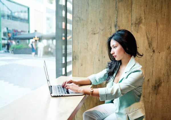 Young businesswoman working from a cafe with a laptop computer. She is dressed in a green blazer.