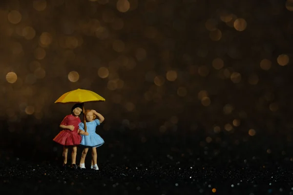 Miniature people toy figure photography. Two girls kid playing outside at rainy day using umbrella at night. Beautifull golden yellow bokeh city light background. Image photo