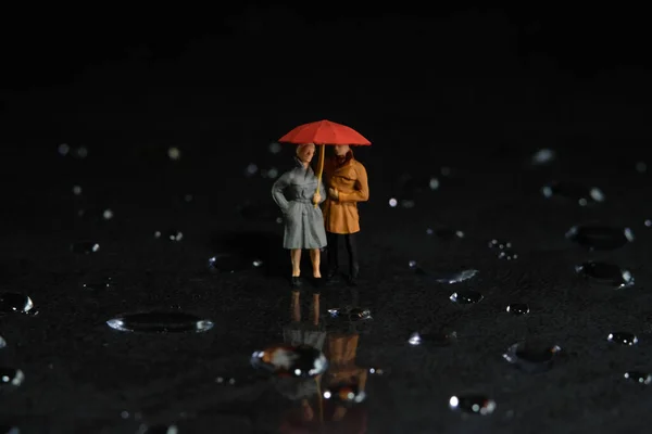 Miniature people toy figure photography. A couple using umbrella, walking on the floor full of droplet. Dark cloudy sky background. Image photo