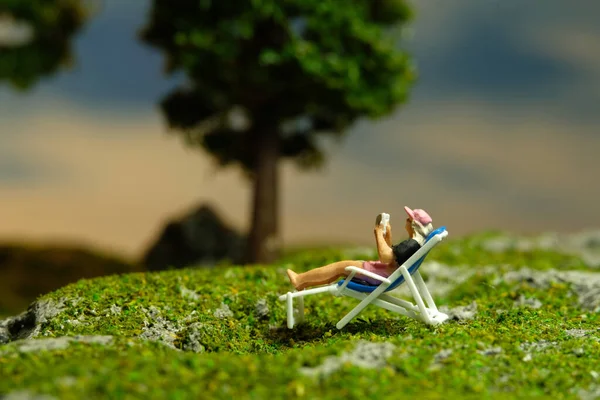 Miniature people toy figure photography. Girl seat on deck chair reading a book, relaxing at garden. Image photo