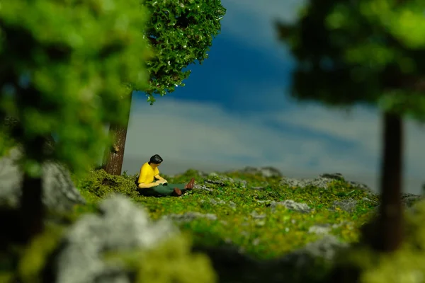 Miniature people toy figure photography. A men sitting under a tree while reading a book, relaxing at garden. Image photo