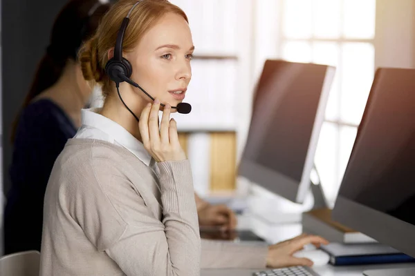 Amicale femme d'affaires caucasienne parlant par casque dans un bureau ensoleillé. Centre d'appels et divers groupes de personnes en affaires — Photo