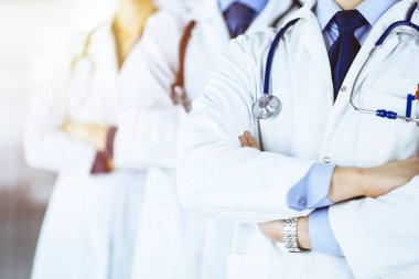 Güneşli bir hastane ofisinde çapraz kolları ve steteskopları olan bir grup modern doktor bir takım olarak ayakta duruyor. Doktorlar hastaları muayene etmeye ve yardım etmeye hazır. Tıbbi yardım, sağlık sigortası, en iyisi.