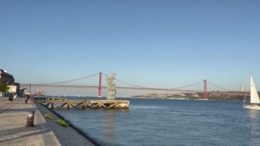 Lisbon,Portugal: July 7,2022- Panoramic view of the iconic The Ponte 25 de Abril bridge over Tagus river shot from Lisbon harbor were people are jogging, relaxing and enjoying the view. 