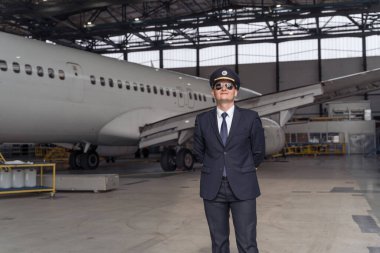 Siyah takım elbiseli erkek pilot bir hangarda duruyor.