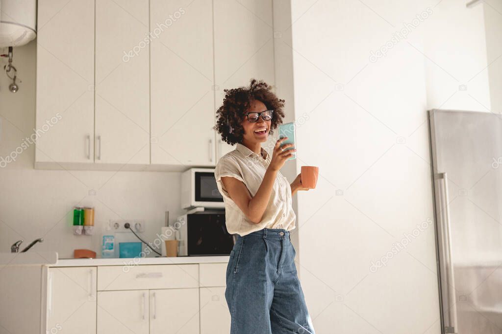 Happy Afro American woman holding phone and cup on the kitchen
