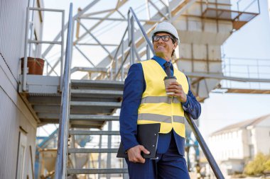 Kahve bardağıyla dışarıda duran neşeli fabrika işçisi.