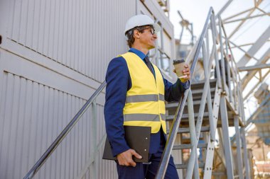 Elinde kahve ve belge tutan erkek fabrika işçisi.