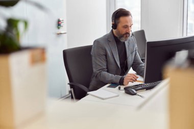 Kulaklıklı mutlu, olgun bir adam açık beyaz bir ofiste oturuyor.