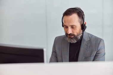 Kulaklıklı mutlu, olgun bir adam açık beyaz bir ofiste oturuyor.