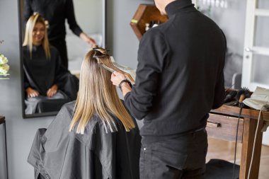 Kendine güvenen erkek stilisti, sarışın beyaz kadın müşterinin saçını boyuyor.