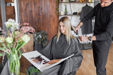 Sarışın bayan müşteri, kuaför saçını keserken kitaptaki renkleri seçiyor.