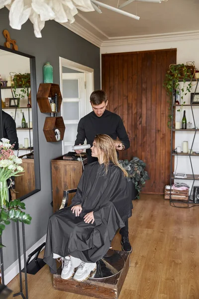 Feliz bela mulher caucasiana cabelo leve no salão de beleza. masculino mestre está secando ela cabelo — Fotografia de Stock